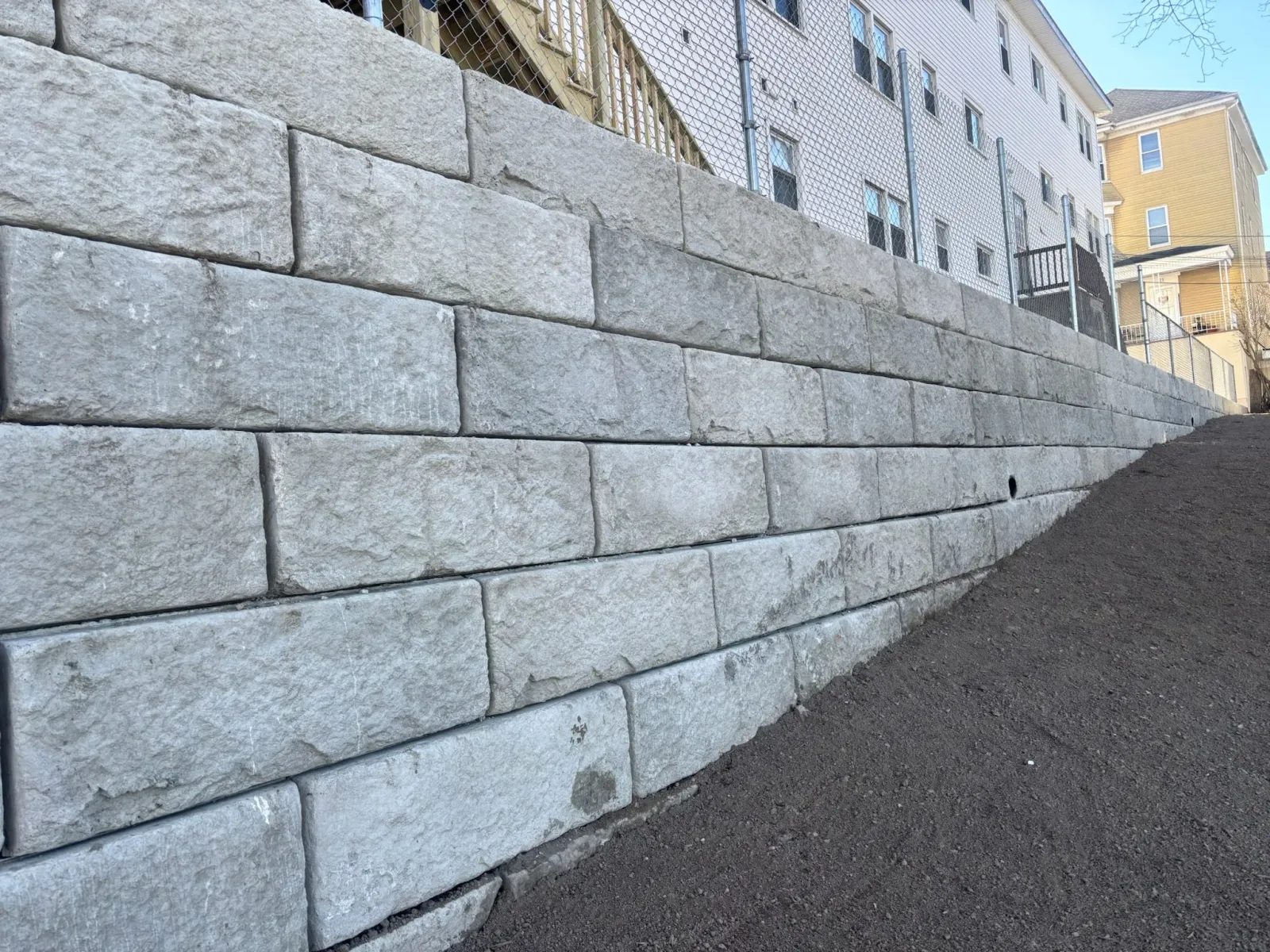 Retaining wall made of concrete blocks with a sloped grassy area and fence on top, adjacent to buildings.