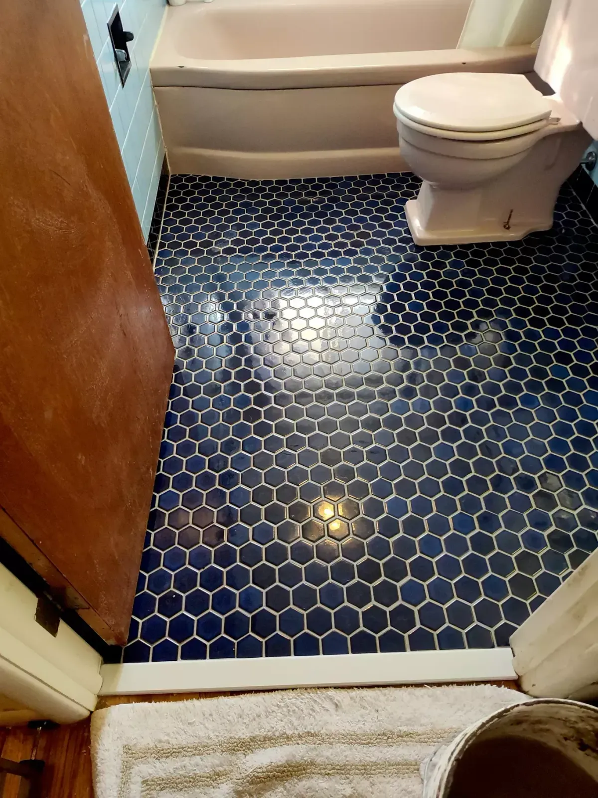 A bathroom with dark blue hexagonal floor tiles, a white toilet, and a bathtub visible in the background.