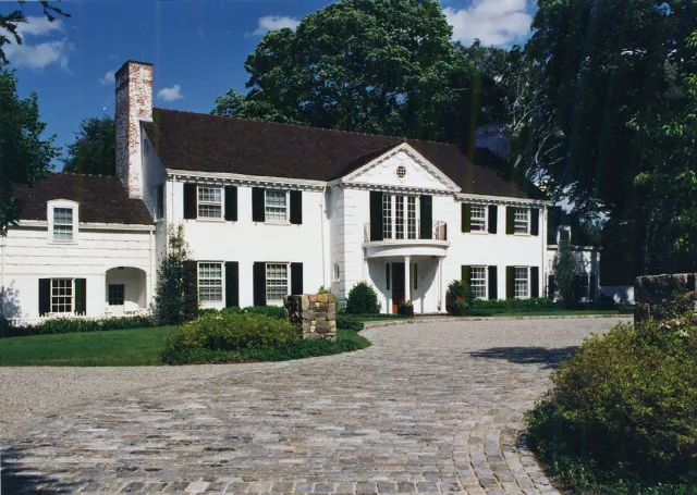 A large white two-story house with black shutters, a red-tiled roof, a chimney, and a small balcony above the front entrance. It is surrounded by a garden with bushes and a stone-paved driveway.