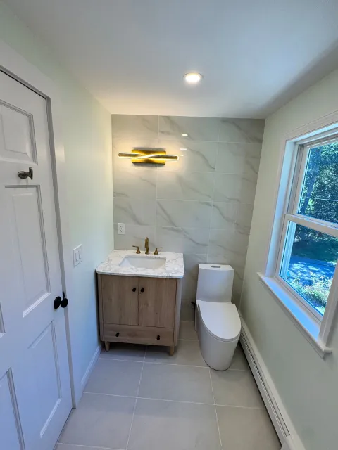 Bathroom with light green walls, wood vanity, marble-like tiled wall, modern toilet, and small window.