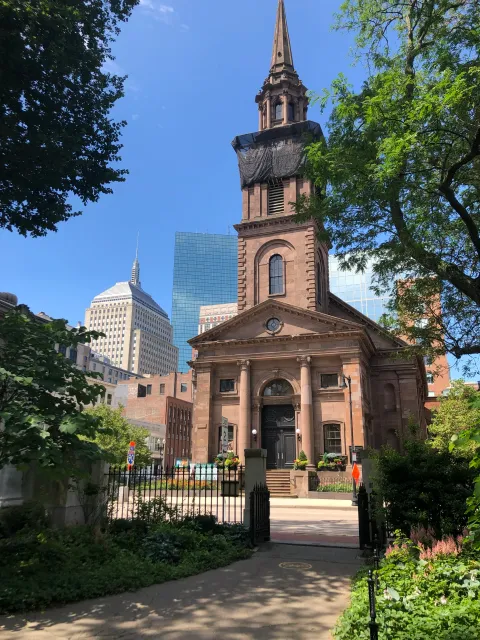 A historic brick church with a covered steeple, surrounded by greenery, set against a modern city skyline on a sunny day.