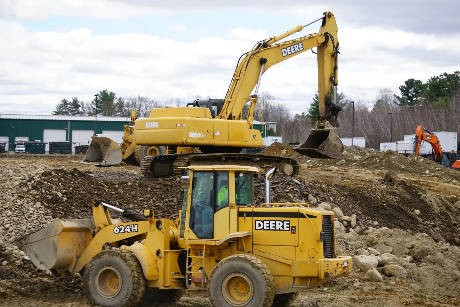 Construction site with yellow John Deere excavator and bulldozer, dirt and rocks on the ground, green warehouse with closed white garage doors in the background, trees, and a cloudy sky.