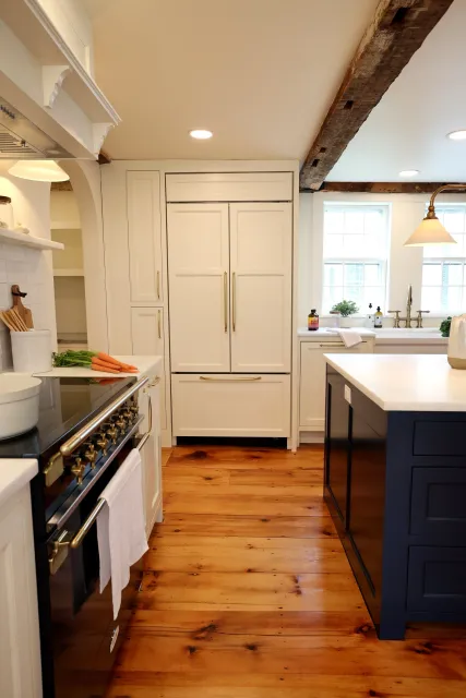 A cozy kitchen with wooden floors, white cabinets with gold handles, a navy blue island, a black stove with gold knobs, and a window above the sink bringing in natural light.