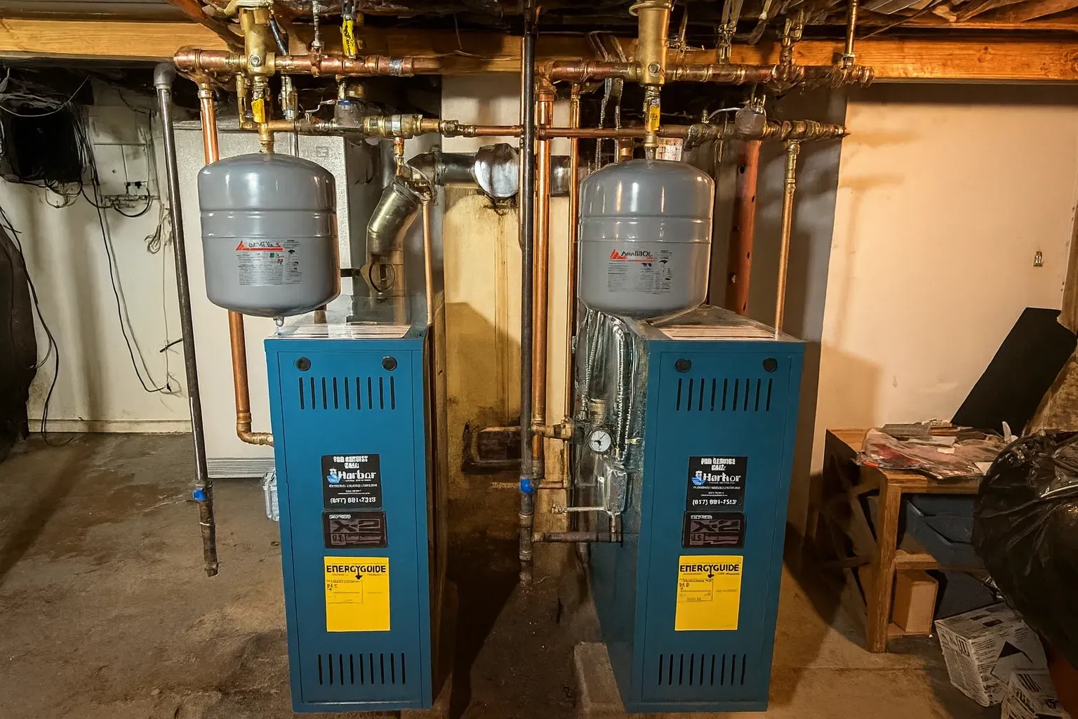 Two blue heating boilers with copper pipes and expansion tanks in a basement setting.
