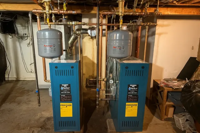 Two blue heating boilers with copper pipes and expansion tanks in a basement setting.