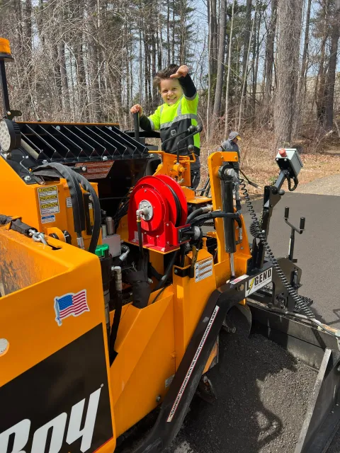 Boy in neon vest on yellow paving machine, fist raised.