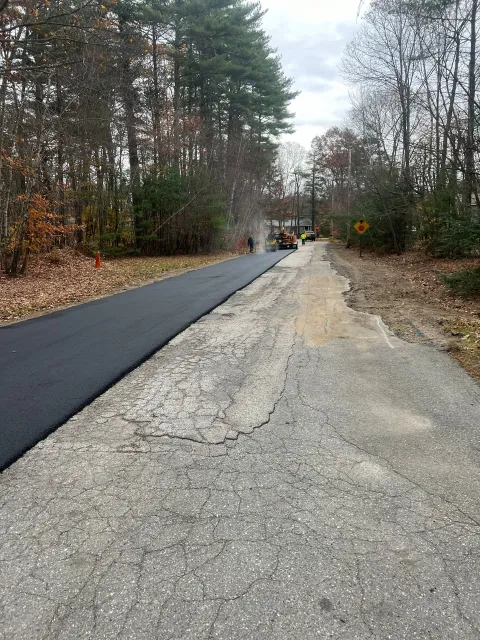 Road paving in progress; asphalt laid on left, older gravel on right, surrounded by trees.