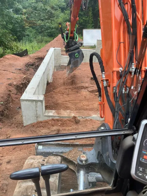 Excavator digging near a concrete foundation in an outdoor setting with red soil.