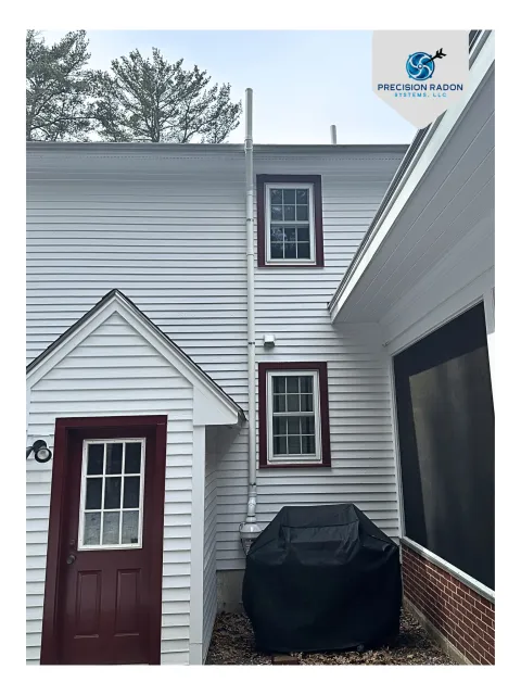 Backyard area with white siding house, maroon door, two windows, a grill covered with a black cover, and a radon mitigation system traveling up the side of the house and venting above the roofline.