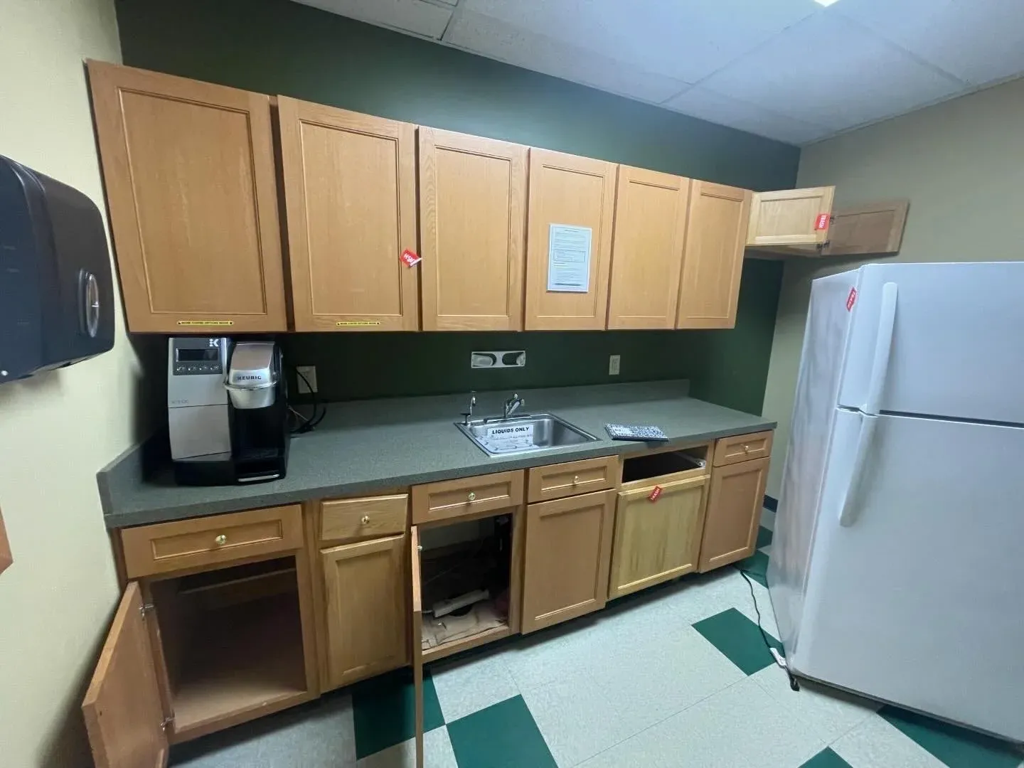 Kitchen area with wooden cabinets, a coffee machine, a refrigerator, and a sink, with a green and white tiled floor.