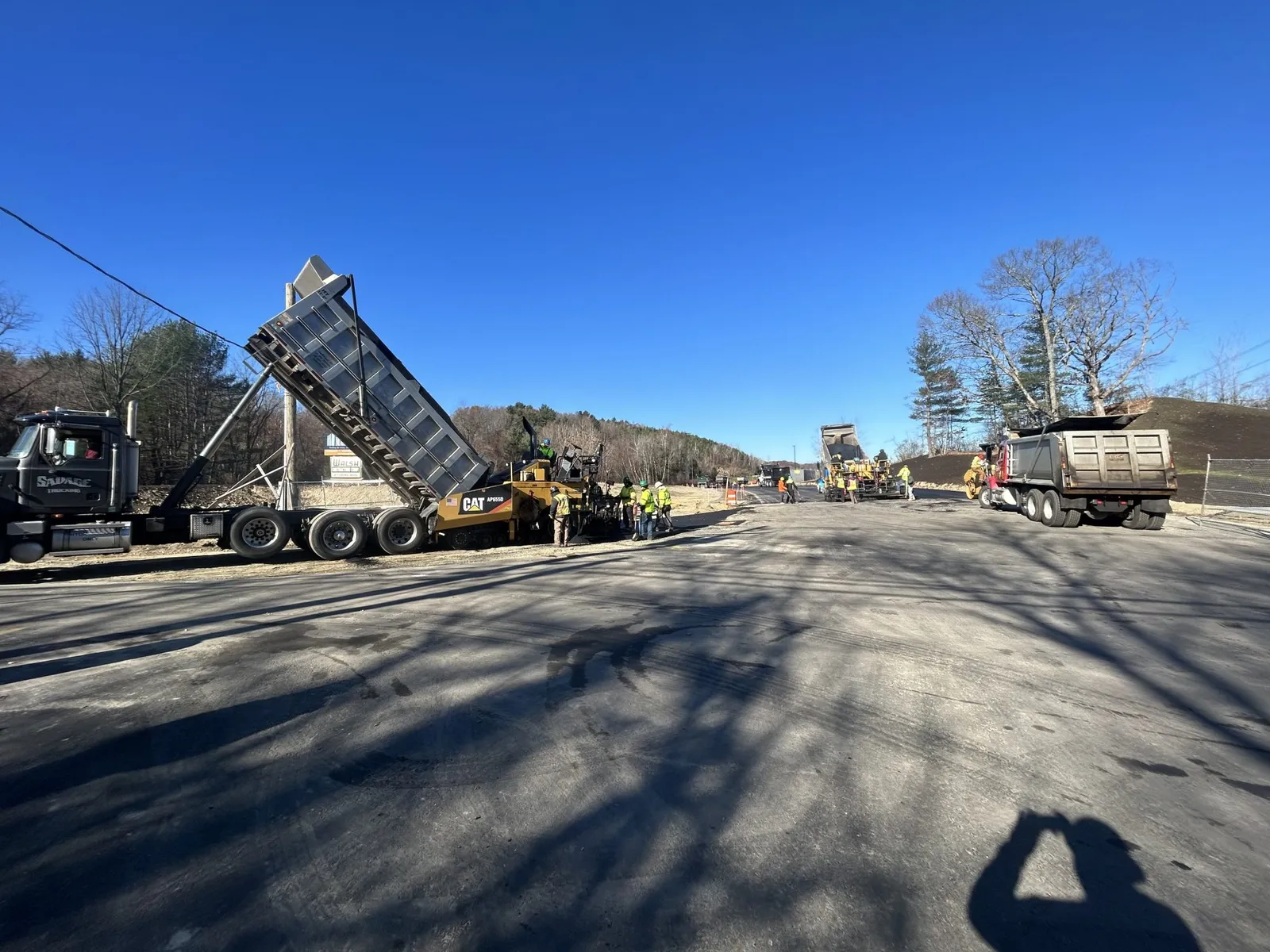 Walsh Contracting Corp paving crew placing hot asphalt with multiple dump trucks on an active commercial paving project in Massachusetts.