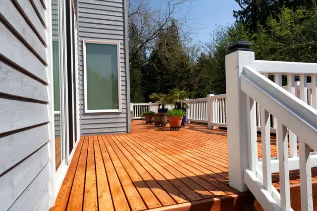 A wooden deck with a curved, light-brown pattern next to a grey house with white railings and potted plants.