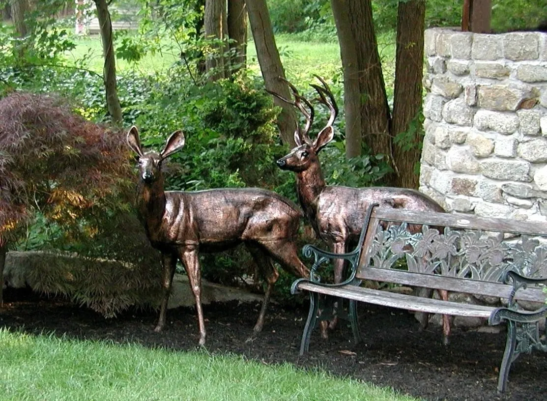 Bronze deer sculptures stand near a stone wall and decorative bench, surrounded by trees and greenery in a tranquil garden setting.