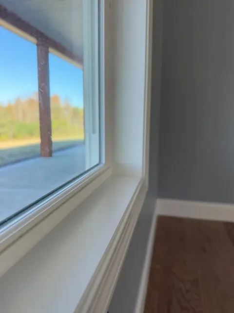 Close-up of a white window frame overlooking a blue sky and trees, with a gray wall and wood floor inside