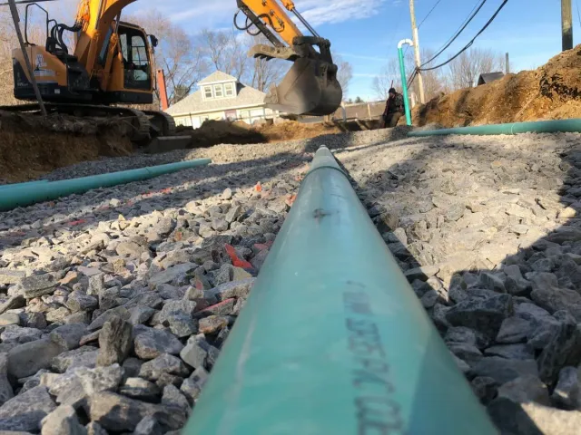 Green pipes in a trench with an excavator in the background, likely a construction site.