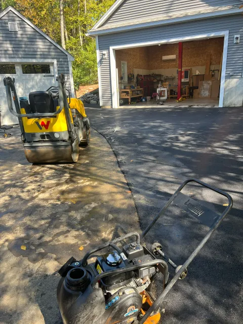 Compactors compacting fresh asphalt on a driveway in front of a garage and small shed.