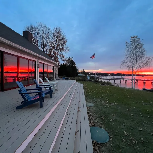 A lakeside scene during sunset with an American flag, a wooden dock, blue and white Adirondack chairs, and a house with large glass doors reflecting the sunset, surrounded by trees and a grassy area.
