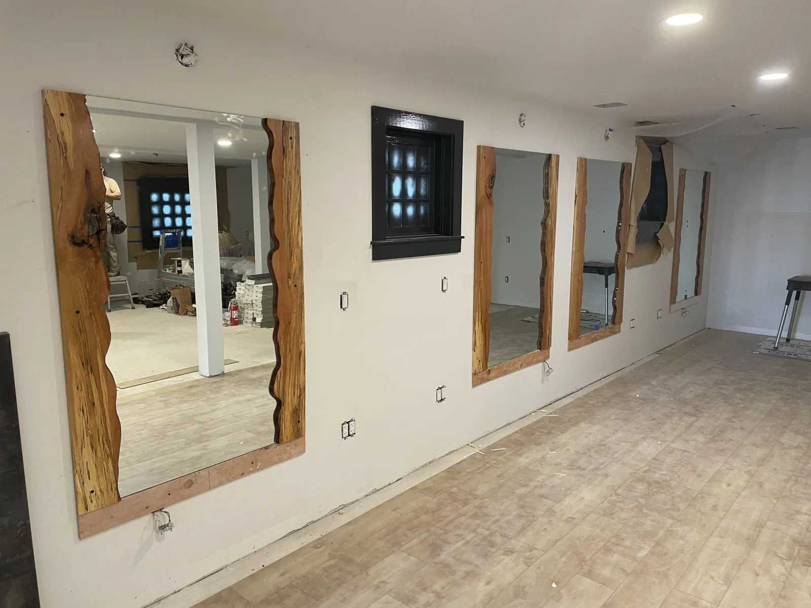 A room under renovation with several large mirrors installed on the wall, each framed with uneven, rustic wood borders. The mirrors are missing their mounting hardware, and the floor is of unfinished wood. There are construction tools and materials v