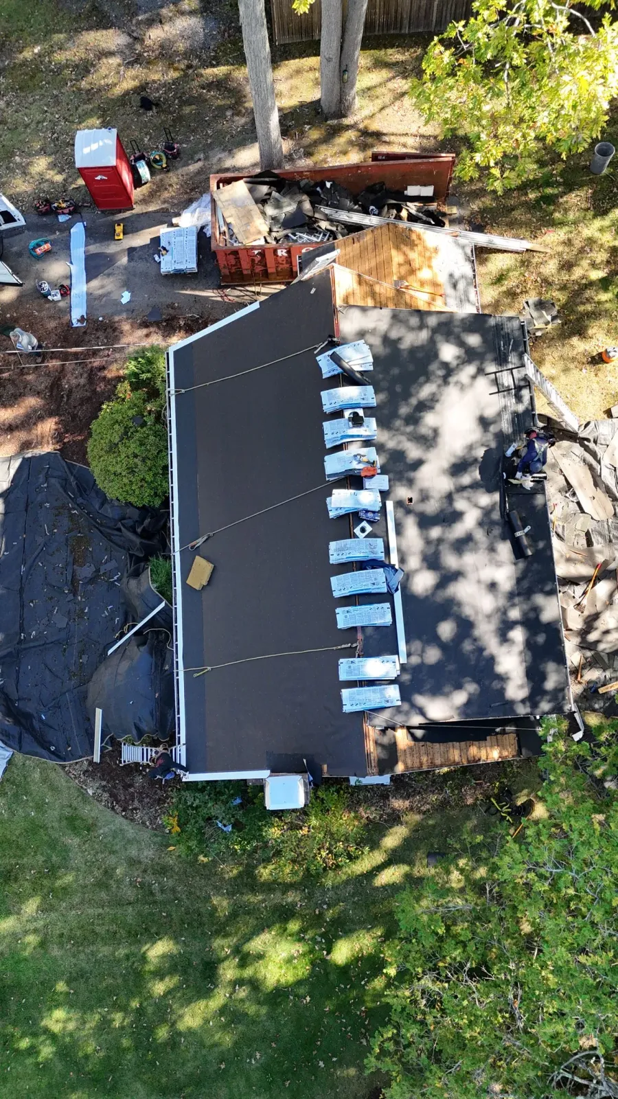 An aerial view of a house roof undergoing repairs, with materials staged along the ridge and debris on the ground below.