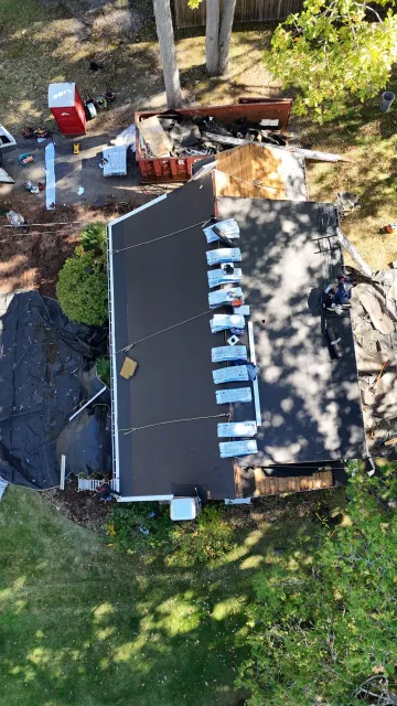 An aerial view of a house roof undergoing repairs, with materials staged along the ridge and debris on the ground below.