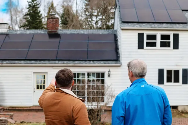 Two people stand in a yard, looking at a white house with solar panels on the roof. One person is pointing towards the panels.