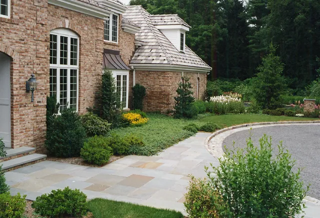 Front yard of a brick house with a curved driveway, stone walkway, landscaped bushes, trees, and garden flowers.