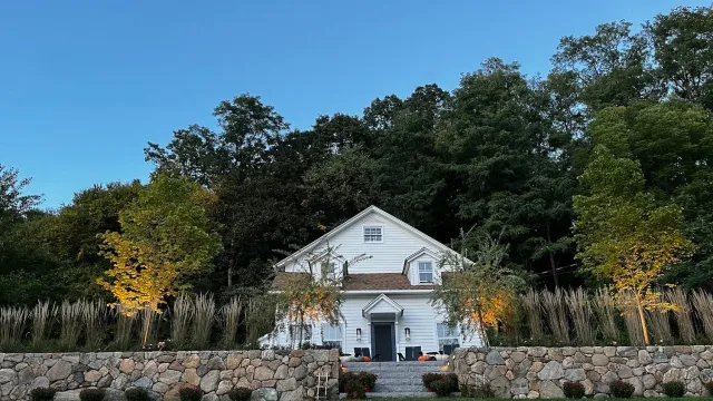 White two-story house with a gabled roof, surrounded by trees and landscaping, stone wall in front, and outdoor seating.