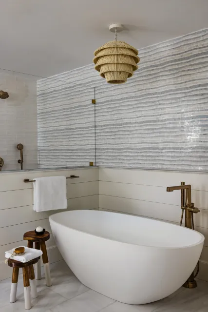 Modern bathroom with a white freestanding bathtub, gold fixtures, a towel hanging on a brass bar, two small wooden stools, a woven pendant light, and a textured mosaic tile wall.