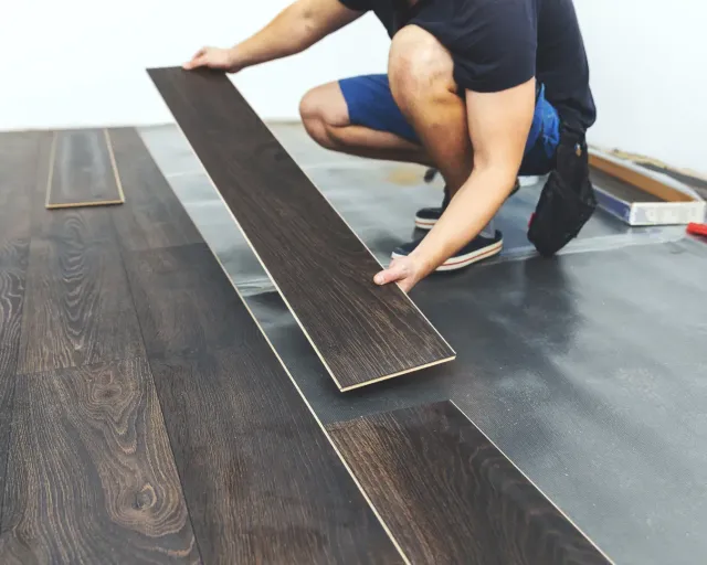 Worker installing dark wood flooring in a room with underlayment sheet visible.