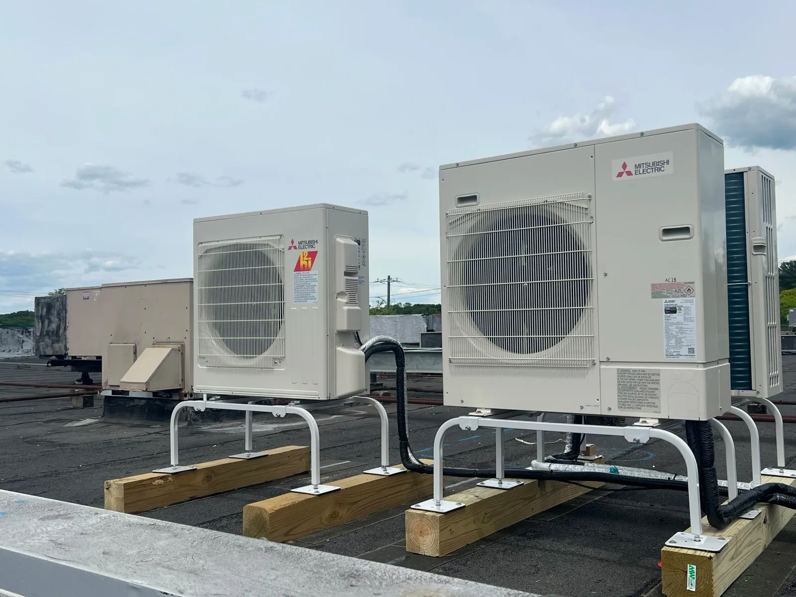 Several large Mitsubishi Electric HVAC units are installed on a rooftop with wooden blocks and metal rails for support, under a partly cloudy sky.