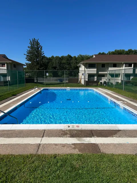 Outdoor swimming pool surrounded by a fence, with apartment buildings in the background and a clear blue sky.