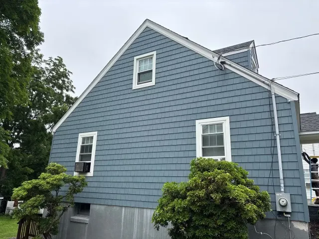 Blue house with white window frames and siding, two windows visible on the upper floors, a tree with green leaves in front, and electrical wires overhead.