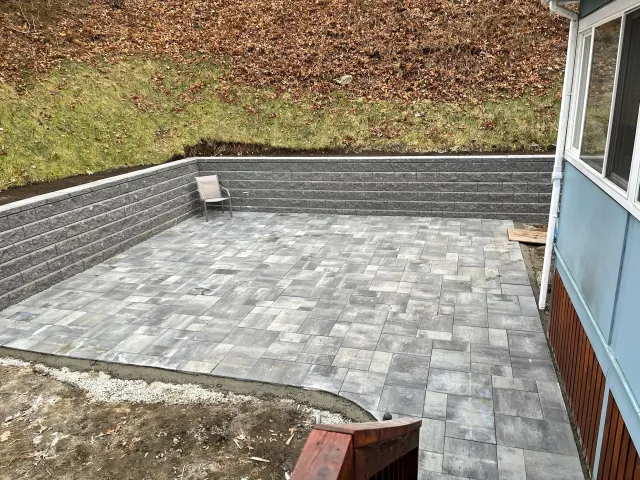 Patio with gray paver stones, wooden bench, gray brick wall, grassy hill with trees and fallen leaves, part of a house with blue siding and white window frames.