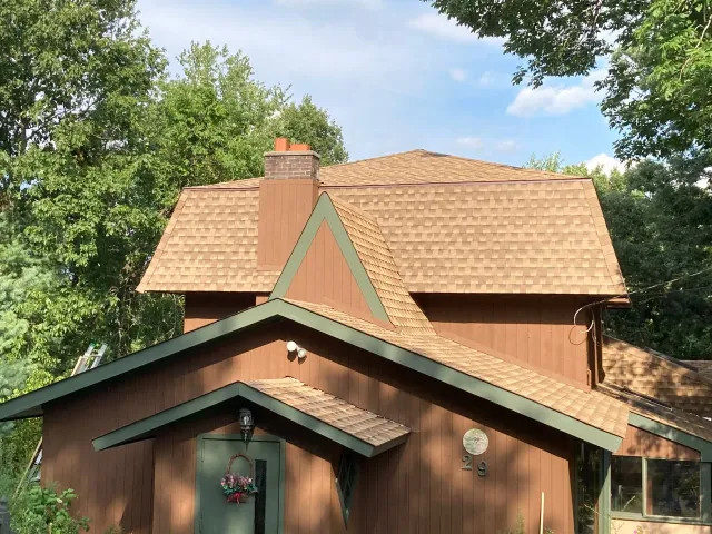 A two-story brown house with a shingled roof, a chimney, and a small front entrance porch, set among green trees.