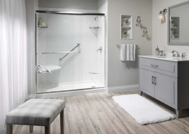 Modern bathroom with a walk-in shower featuring white subway tile walls, chrome grab bars, a hexagonal corner seat, and a clear glass sliding door. Room includes a gray vanity, light wood flooring, and decorative wall shelving.