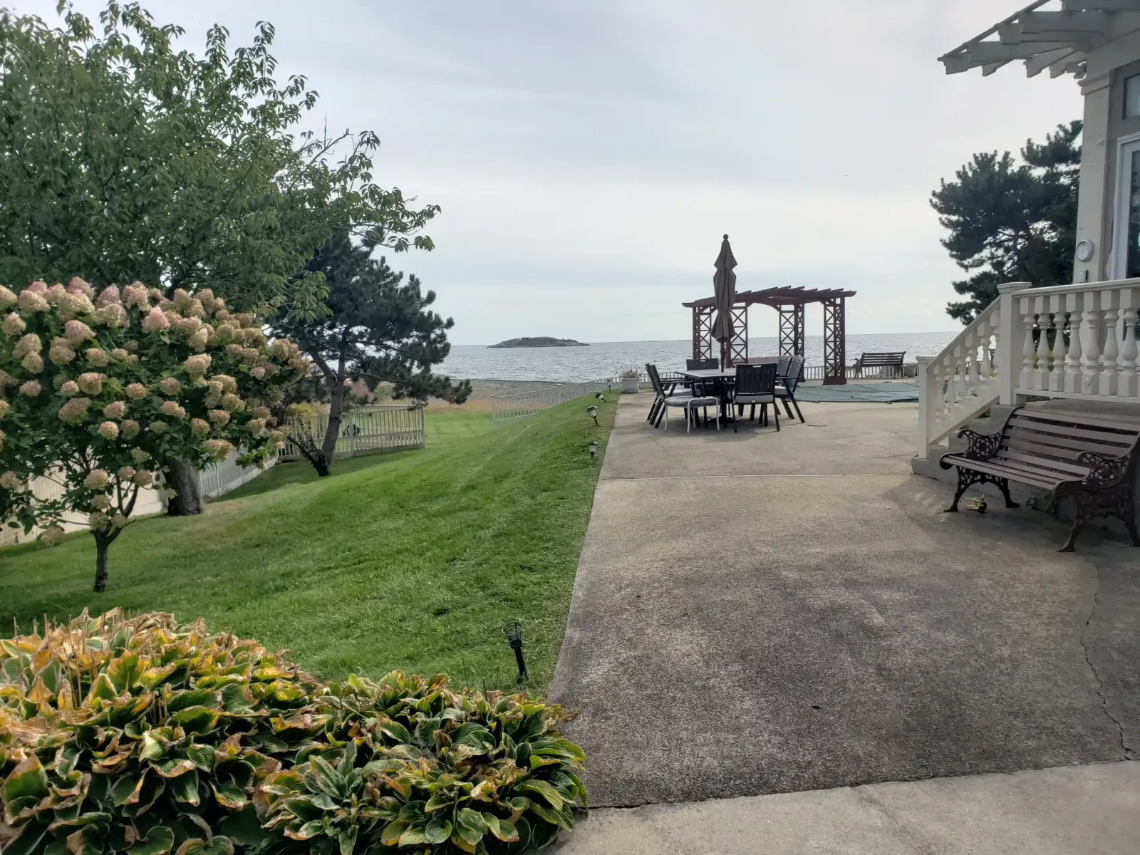 Sloped green lawn leads to a gray ocean view, with pale hydrangeas on the left and patio chairs under a pergola by the shore