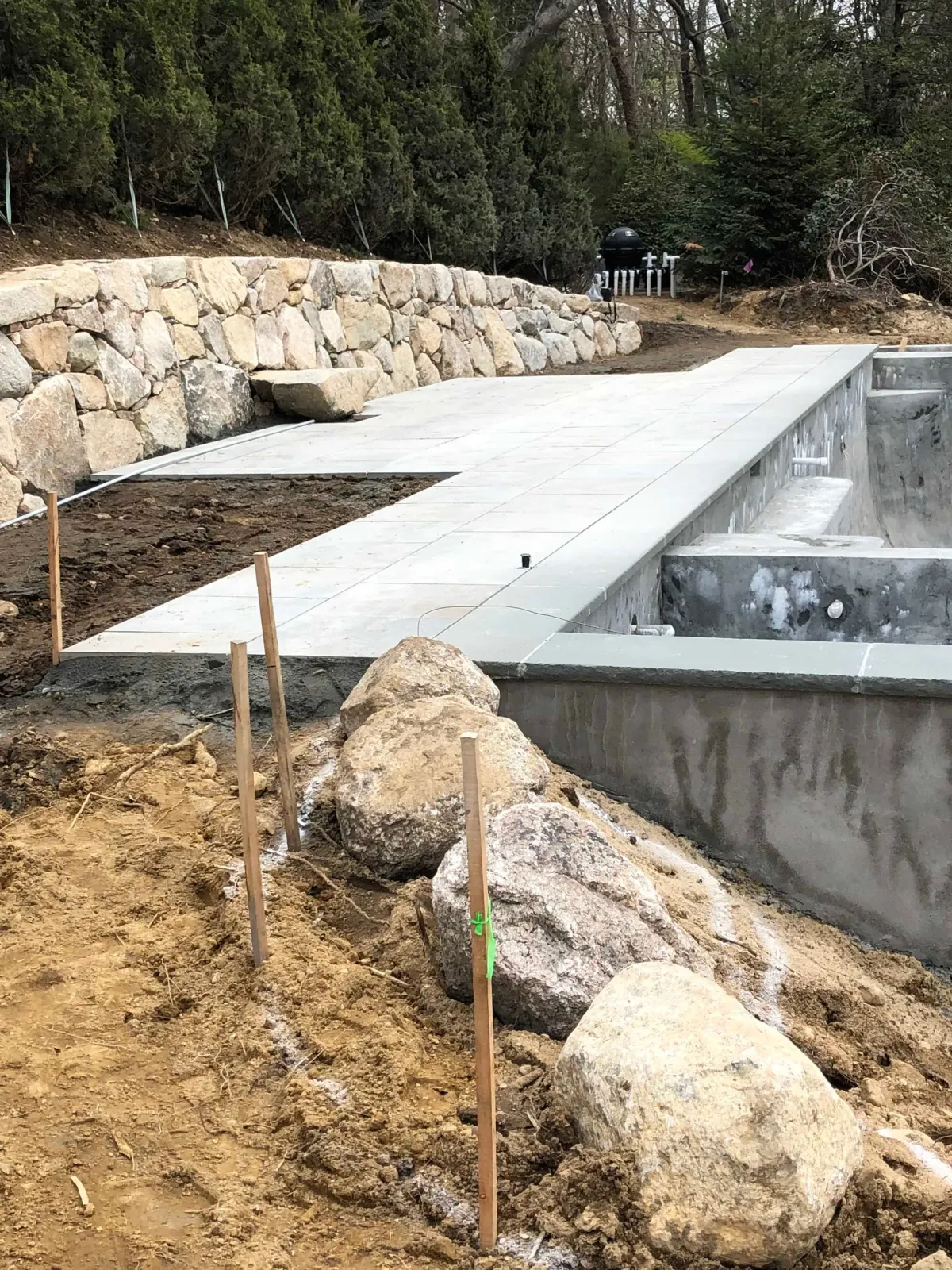 A construction site featuring a concrete pool frame, stone retaining walls, and a new patio area under development.