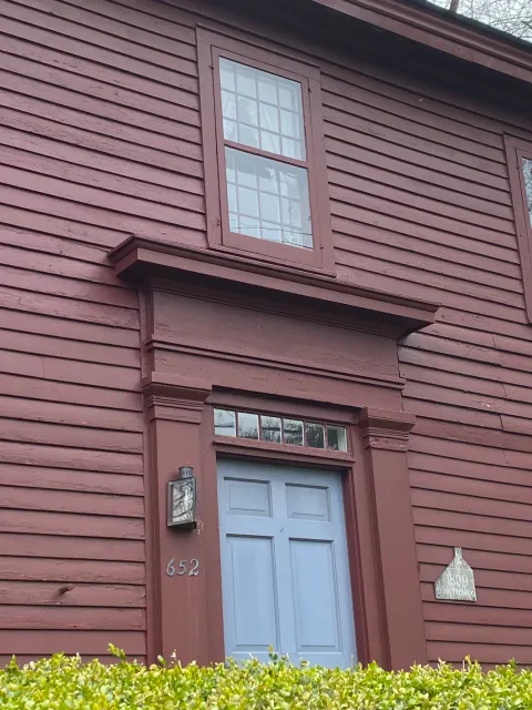 Close-up of a house with burgundy siding, blue front door, and two multi-pane windows, one above the door and one above. The house number 652 is displayed near the door, and a light fixture is mounted beside it. A hedge is in the foreground.