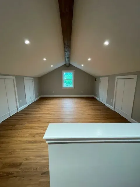 Empty attic room with wood floor, white walls, recessed lights, and a single small window.