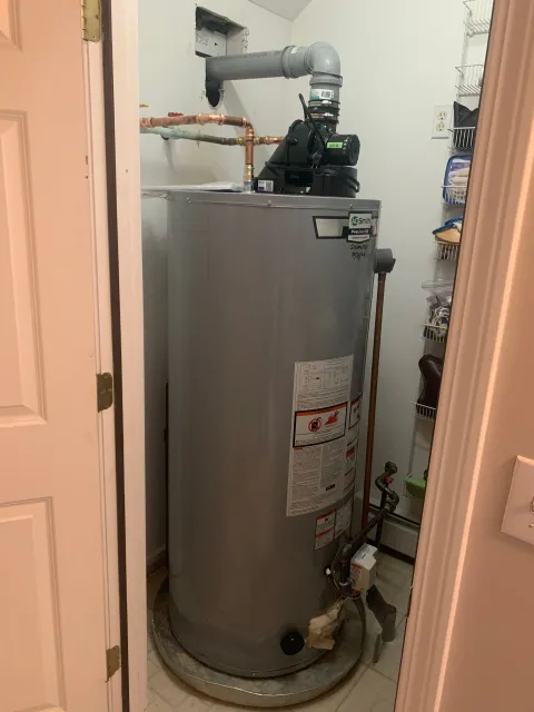 A tall, silver water heater in a small utility closet, with pipes and a pump on top.