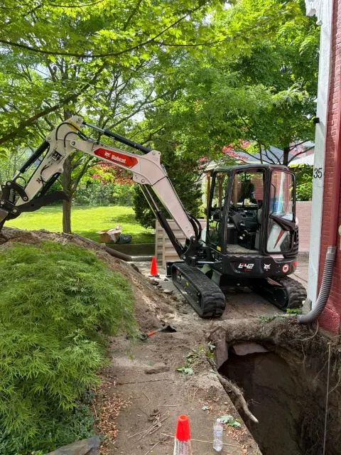 Excavator working beside a trench on a wooded path, with traffic cones marking the excavation area.