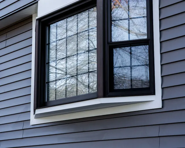 Gray house siding with a bay window and white trim, reflecting bare tree branches.