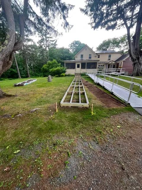 A house with raised garden beds in a grassy yard, framed by trees and a gravel path.