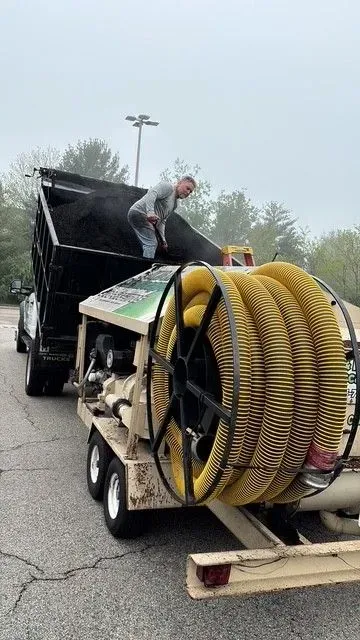A man unloading material from a dump truck into a large machine with a yellow hose reel, outdoors on a cloudy day.