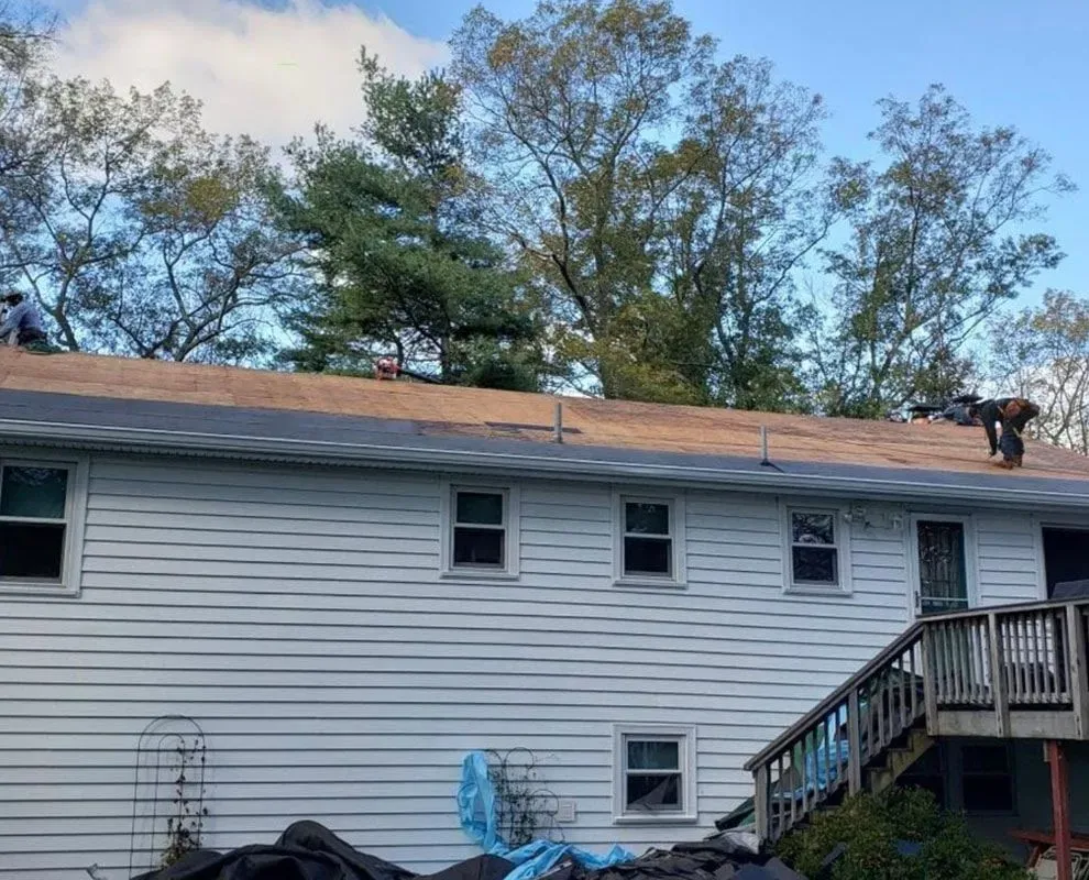 Workers on a rooftop, installing shingles. White siding, trees in background, and a wooden deck.