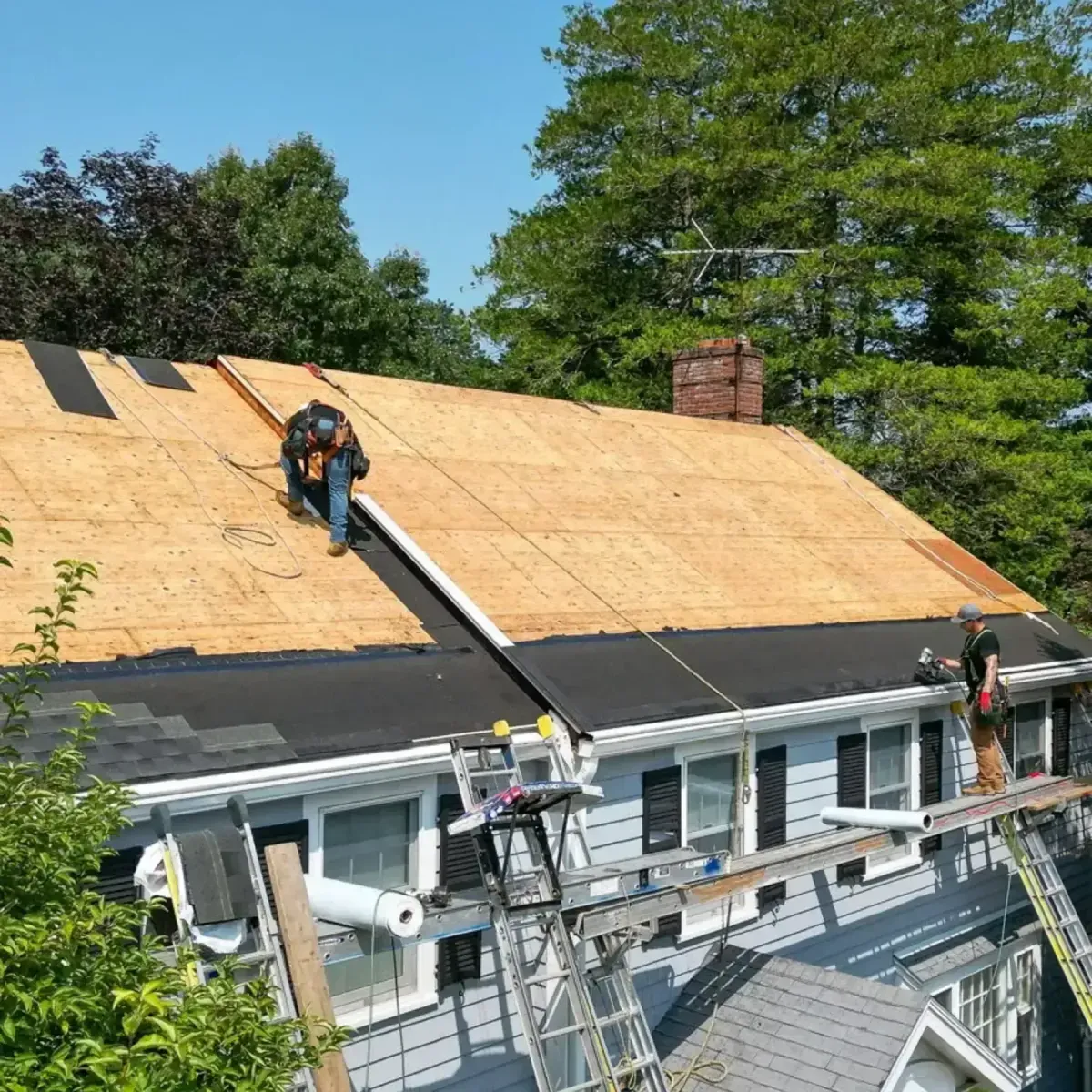 Roofing crew working on a residential roof installation, featuring plywood sheathing and black underlayment, with scaffolding and tools visible, showcasing commercial roofing services by Crown Roofing.