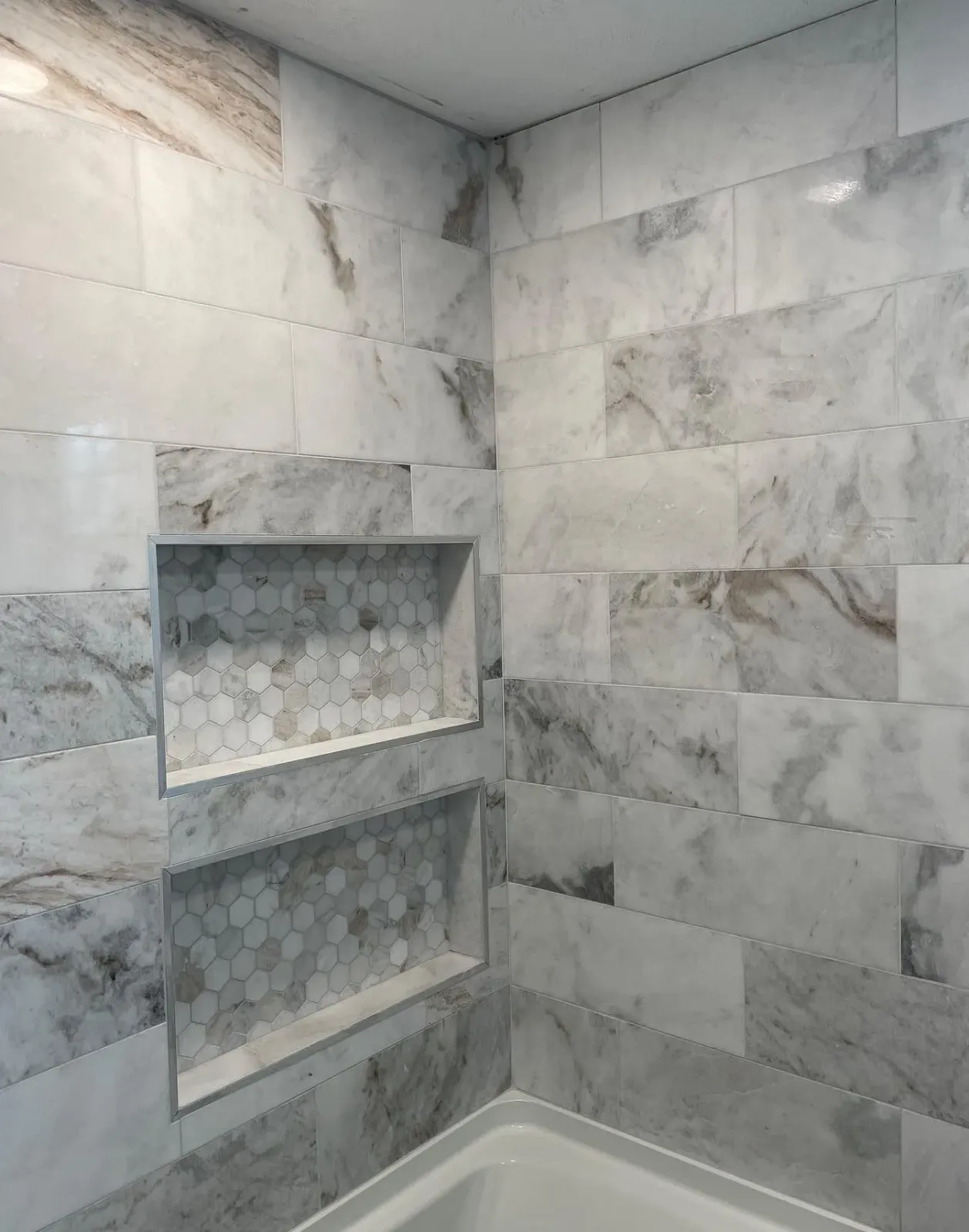 A corner of a tiled shower with two built-in storage niches featuring a gray and white marble pattern throughout.