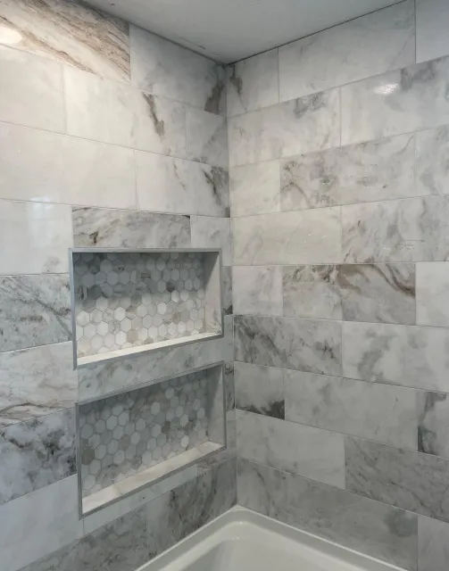 A corner of a tiled shower with two built-in storage niches featuring a gray and white marble pattern throughout.