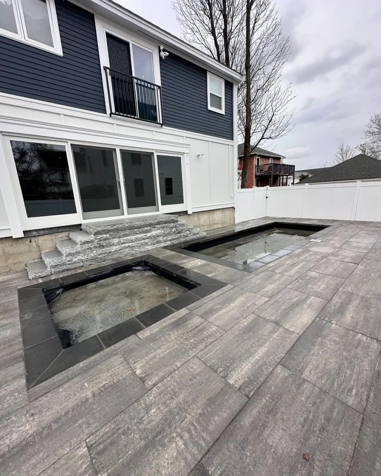 Backyard patio with two rectangular in-ground pools and a two-story blue and white house. Gray pavers and steps.