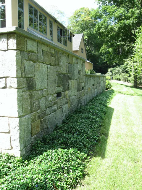 Backyard view with manicured lawn, stone retaining wall, patio seating area, and mature trees providing natural shade and privacy.